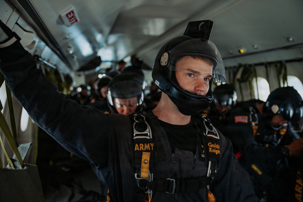 A group of U.S. Army Golden Knights parachute team members are seated inside a military aircraft, fully equipped with skydiving helmets, harnesses, and jumpsuits. The foreground subject is a young male soldier wearing a black helmet with a mounted camera, a parachute harness labeled 'ARMY' and 'Golden Knights'. The team appears to be preparing for a jump, with multiple personnel visible in the background.