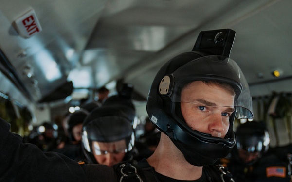 A group of U.S. Army Golden Knights parachute team members are seated inside a military aircraft, fully equipped with skydiving helmets, harnesses, and jumpsuits. The foreground subject is a young male soldier wearing a black helmet with a mounted camera, a parachute harness labeled 'ARMY' and 'Golden Knights'. The team appears to be preparing for a jump, with multiple personnel visible in the background.