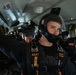 A group of U.S. Army Golden Knights parachute team members are seated inside a military aircraft, fully equipped with skydiving helmets, harnesses, and jumpsuits. The foreground subject is a young male soldier wearing a black helmet with a mounted camera, a parachute harness labeled 'ARMY' and 'Golden Knights'. The team appears to be preparing for a jump, with multiple personnel visible in the background.