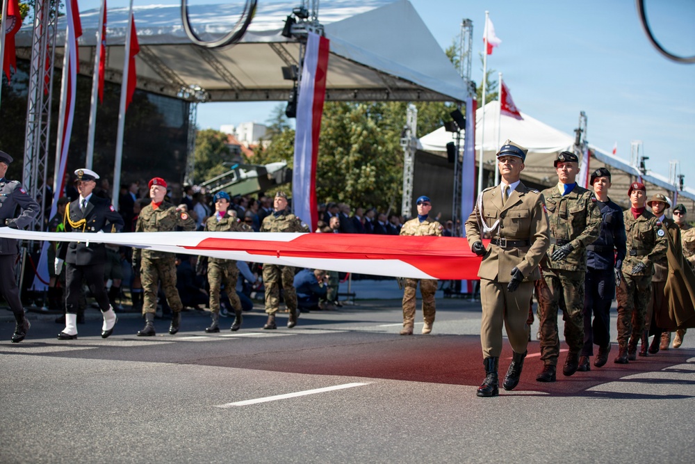 A military parade is taking place on a paved street, with soldiers in various uniforms carrying a large red-and-white flag horizontally. The flag's colors — white and red — correspond to the Polish national flag, and additional Polish flags are visible on poles in the background. A stage structure with lighting rigs and a crowd of spectators is visible behind the marching formation.