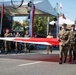 A military parade is taking place on a paved street, with soldiers in various uniforms carrying a large red-and-white flag horizontally. The flag's colors — white and red — correspond to the Polish national flag, and additional Polish flags are visible on poles in the background. A stage structure with lighting rigs and a crowd of spectators is visible behind the marching formation.