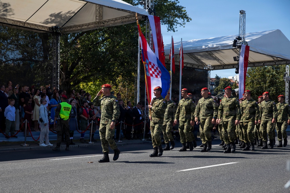 A military parade is taking place on a paved street, featuring soldiers in green camouflage uniforms and distinctive red berets marching in formation. A prominent Croatian flag (red-white-blue with the Croatian coat of arms checkerboard) is being carried by one of the soldiers. Spectators line the left side of the route behind a red rope barrier, and white canopy tents serve as viewing platforms or official stands.