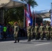 A military parade is taking place on a paved street, featuring soldiers in green camouflage uniforms and distinctive red berets marching in formation. A prominent Croatian flag (red-white-blue with the Croatian coat of arms checkerboard) is being carried by one of the soldiers. Spectators line the left side of the route behind a red rope barrier, and white canopy tents serve as viewing platforms or official stands.