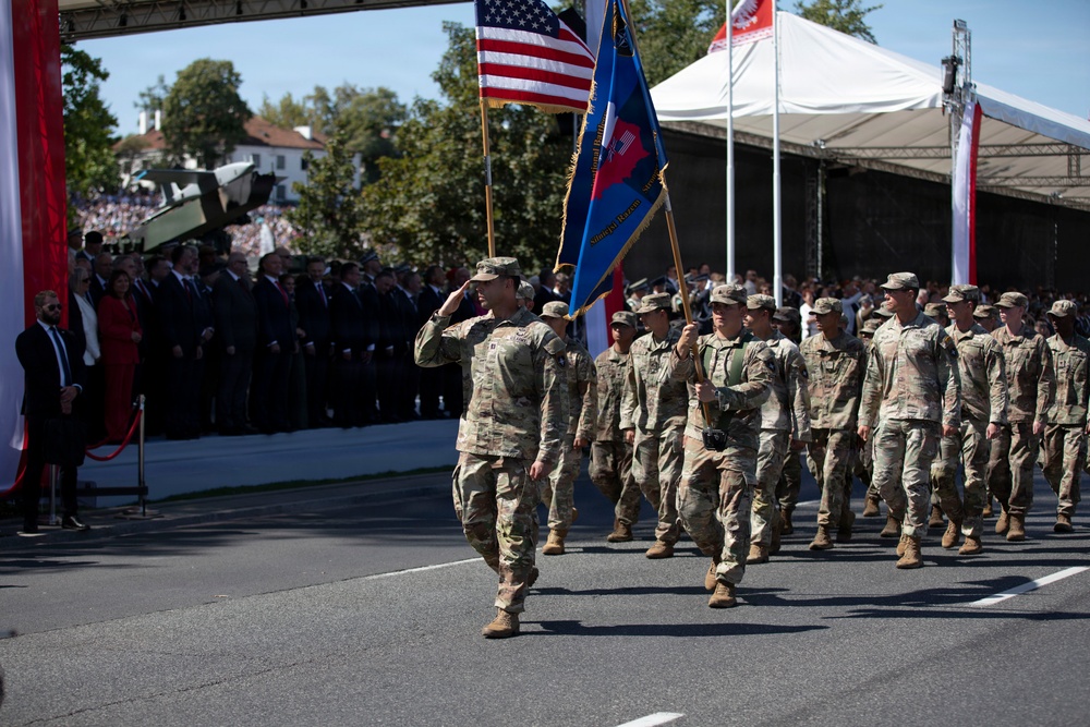 A formation of U.S. Army soldiers in OCP (Operational Camouflage Pattern) uniforms marches in a military parade, carrying the American flag and a unit guidon/colors. A lead soldier renders a salute toward a reviewing stand where dignitaries and officials are gathered. The event appears to be a formal military parade, likely held in Poland based on the red-and-white national colors visible on flags and banners.