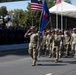 A formation of U.S. Army soldiers in OCP (Operational Camouflage Pattern) uniforms marches in a military parade, carrying the American flag and a unit guidon/colors. A lead soldier renders a salute toward a reviewing stand where dignitaries and officials are gathered. The event appears to be a formal military parade, likely held in Poland based on the red-and-white national colors visible on flags and banners.