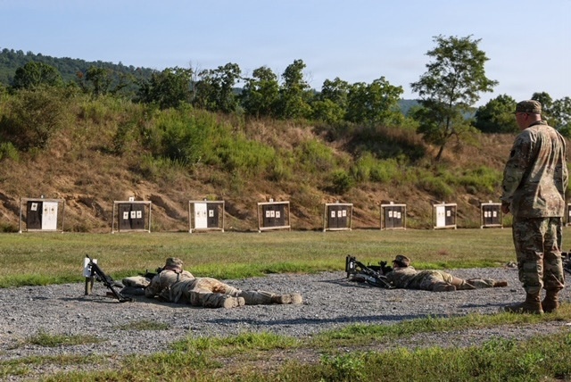 The image shows a military rifle range with two soldiers in prone firing positions aiming downrange at a series of paper targets mounted on wooden frames. A third soldier, likely a range safety officer or instructor, stands to the right observing the shooters. The setting appears to be an outdoor training range with a natural earthen berm backstop and wooded hillside in the background.