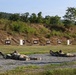 The image shows a military rifle range with two soldiers in prone firing positions aiming downrange at a series of paper targets mounted on wooden frames. A third soldier, likely a range safety officer or instructor, stands to the right observing the shooters. The setting appears to be an outdoor training range with a natural earthen berm backstop and wooded hillside in the background.