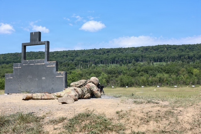 A soldier in full camouflage uniform and combat gear is in a prone firing position on a military shooting range. The soldier is aiming a rifle downrange toward distant targets visible in the background. A large concrete shooting position barrier or target structure is visible to the left.