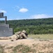 A soldier in full camouflage uniform and combat gear is in a prone firing position on a military shooting range. The soldier is aiming a rifle downrange toward distant targets visible in the background. A large concrete shooting position barrier or target structure is visible to the left.