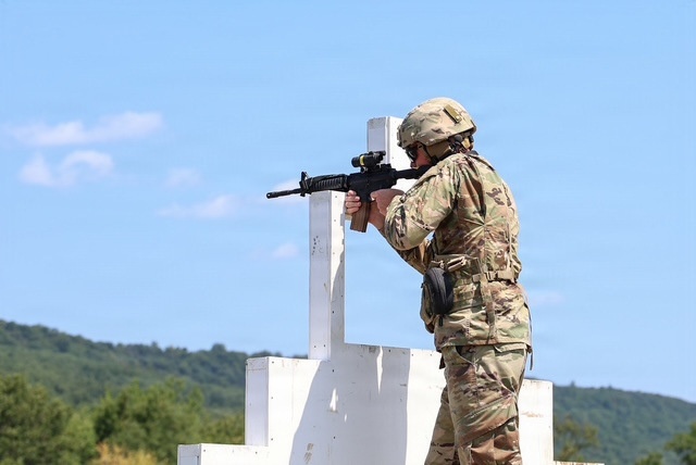 A soldier in full military gear is positioned behind a white barricade structure, aiming a rifle downrange at what appears to be a military shooting range. The soldier is wearing MultiCam (OCP) camouflage uniform and a tactical helmet. The setting appears to be an outdoor range during daytime with clear skies and forested hills in the background.