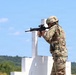 A soldier in full military gear is positioned behind a white barricade structure, aiming a rifle downrange at what appears to be a military shooting range. The soldier is wearing MultiCam (OCP) camouflage uniform and a tactical helmet. The setting appears to be an outdoor range during daytime with clear skies and forested hills in the background.