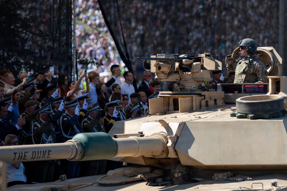 A military parade featuring an M1 Abrams main battle tank named 'The Dune' passes before a large crowd of spectators and uniformed military officers. A soldier in combat gear and helmet is visible atop the tank, appearing to salute. The event appears to be a formal military ceremony or national parade with both civilian and military dignitaries present.