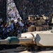 A military parade featuring an M1 Abrams main battle tank named 'The Dune' passes before a large crowd of spectators and uniformed military officers. A soldier in combat gear and helmet is visible atop the tank, appearing to salute. The event appears to be a formal military ceremony or national parade with both civilian and military dignitaries present.