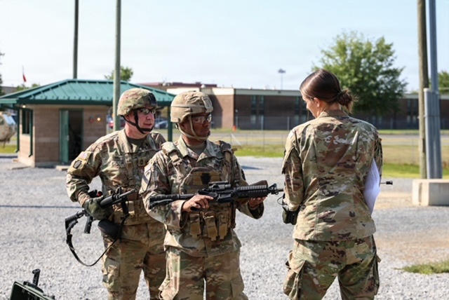 Three military personnel in U.S. Army OCP (Operational Camouflage Pattern) uniforms are engaged in what appears to be a briefing or inspection at a military installation. Two soldiers wearing helmets and carrying M4-style rifles are facing a third individual whose back is to the camera. The setting appears to be an outdoor area of a military base with buildings and a gravel surface visible in the background.