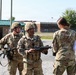 Three military personnel in U.S. Army OCP (Operational Camouflage Pattern) uniforms are engaged in what appears to be a briefing or inspection at a military installation. Two soldiers wearing helmets and carrying M4-style rifles are facing a third individual whose back is to the camera. The setting appears to be an outdoor area of a military base with buildings and a gravel surface visible in the background.