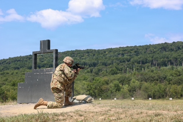 A soldier in full camouflage uniform and helmet is kneeling in a firing position on a military shooting range, aiming a rifle from behind a large metal barricade structure. The setting appears to be an outdoor live-fire training range with multiple white target markers visible downrange. The background features a wooded hillside under a partly cloudy blue sky.