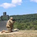 A soldier in full camouflage uniform and helmet is kneeling in a firing position on a military shooting range, aiming a rifle from behind a large metal barricade structure. The setting appears to be an outdoor live-fire training range with multiple white target markers visible downrange. The background features a wooded hillside under a partly cloudy blue sky.