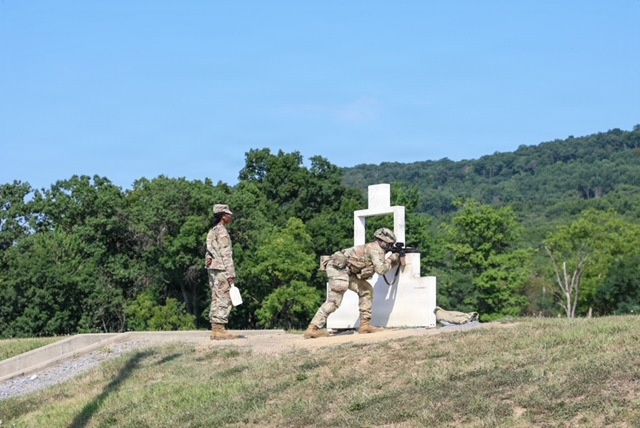 The image shows military personnel conducting a weapons qualification or marksmanship training exercise at an outdoor firing range. Two soldiers are positioned at a white concrete shooting barricade structure, with one in a firing stance aiming a rifle, while a third soldier stands nearby observing and holding what appears to be a clipboard or paperwork. The setting is a grassy range area with dense tree-covered hills in the background under a clear blue sky.