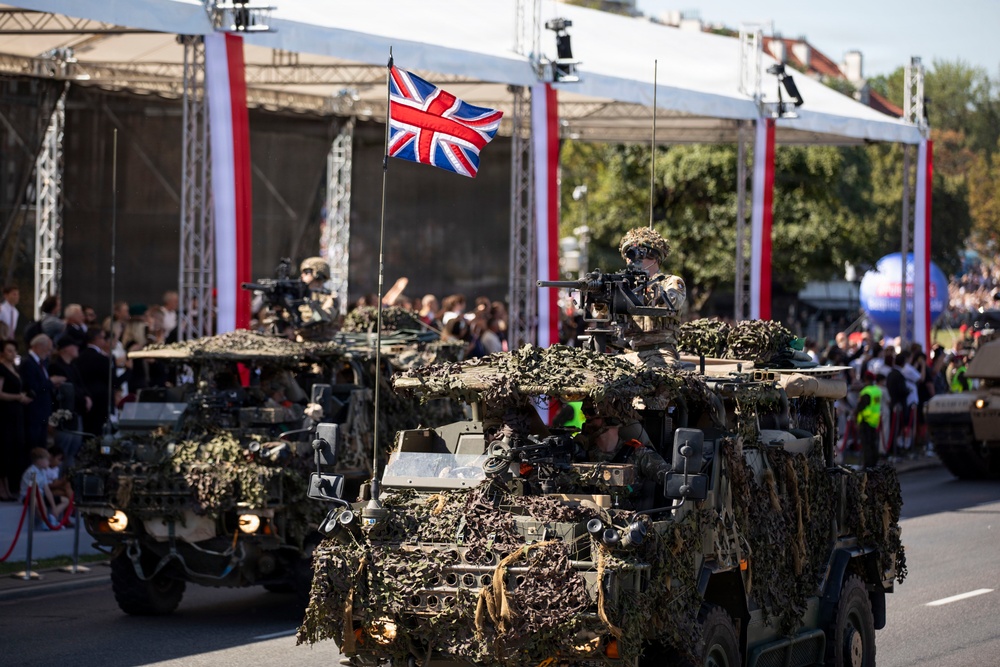 A military parade featuring British Army vehicles heavily draped in camouflage netting moves along a street in front of a grandstand with red and white decorative banners. The lead vehicle displays a Union Jack flag and has armed soldiers manning weapon stations on top. Crowds of spectators and dignitaries line the parade route behind barriers.