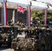 A military parade featuring British Army vehicles heavily draped in camouflage netting moves along a street in front of a grandstand with red and white decorative banners. The lead vehicle displays a Union Jack flag and has armed soldiers manning weapon stations on top. Crowds of spectators and dignitaries line the parade route behind barriers.