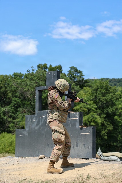 A U.S. Army soldier in full Operational Camouflage Pattern (OCP) uniform is conducting a weapons qualification or training exercise at an outdoor range. The soldier is in a standing shooting stance, aiming a rifle with an optic attachment toward an unseen target. The setting appears to be a military training facility with constructed barrier structures and a wooded background.