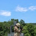 A U.S. Army soldier in full Operational Camouflage Pattern (OCP) uniform is conducting a weapons qualification or training exercise at an outdoor range. The soldier is in a standing shooting stance, aiming a rifle with an optic attachment toward an unseen target. The setting appears to be a military training facility with constructed barrier structures and a wooded background.