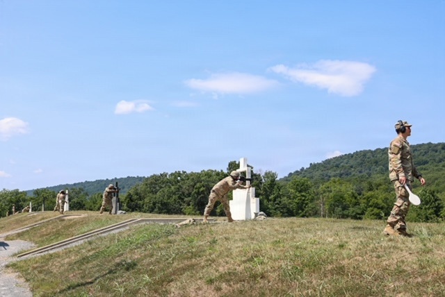 The image shows military personnel in camouflage uniforms conducting what appears to be a rifle range or weapons qualification exercise at an outdoor training facility. Several soldiers are positioned along a berm or firing line, with at least one appearing to aim a weapon at target structures. A range safety officer or instructor is visible in the foreground holding what appears to be a scoring paddle or range control device.