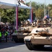 A military parade featuring M1 Abrams main battle tanks moving along a paved street past a reviewing stand with spectators. The tanks display unit markings consistent with U.S. Army designation '1A 2-37' (2nd Battalion, 37th Armor Regiment). Red and white decorative elements on the stage structure suggest this may be a Polish national event, likely Polish Armed Forces Day.
