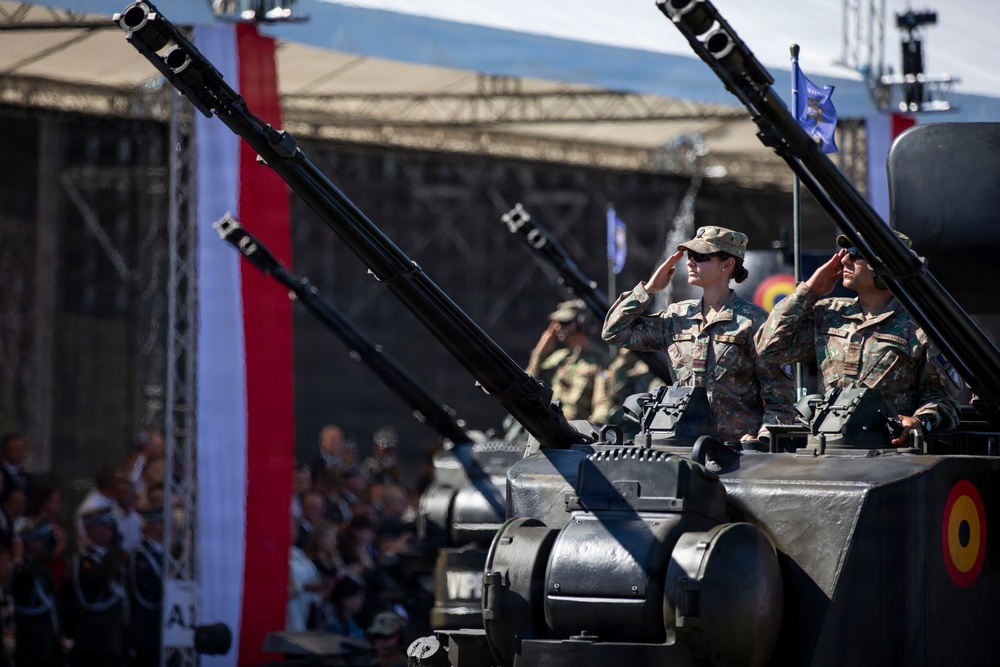 A military parade featuring armored vehicles with anti-aircraft gun systems, with soldiers in camouflage uniforms saluting from atop the vehicles. A large crowd of spectators is visible in the background alongside red and white vertical banners. The scene appears to be a formal national military ceremony or independence day parade.