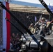 A military parade featuring armored vehicles with anti-aircraft gun systems, with soldiers in camouflage uniforms saluting from atop the vehicles. A large crowd of spectators is visible in the background alongside red and white vertical banners. The scene appears to be a formal national military ceremony or independence day parade.