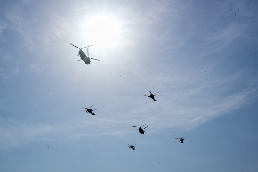 A formation of military helicopters flying in a blue, partially cloudy sky with the sun visible in the upper portion of the frame. One large tandem-rotor helicopter (consistent with a CH-47 Chinook type) is prominently silhouetted near the sun, while five smaller helicopters (consistent with attack or utility helicopter types) fly in a loose formation below. All aircraft are rendered as dark silhouettes due to backlighting from the bright sun.