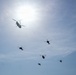 A formation of military helicopters flying in a blue, partially cloudy sky with the sun visible in the upper portion of the frame. One large tandem-rotor helicopter (consistent with a CH-47 Chinook type) is prominently silhouetted near the sun, while five smaller helicopters (consistent with attack or utility helicopter types) fly in a loose formation below. All aircraft are rendered as dark silhouettes due to backlighting from the bright sun.
