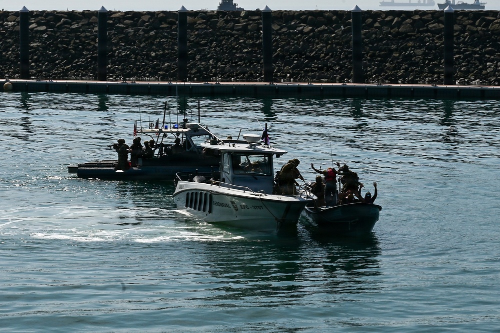 The image shows a maritime law enforcement or military interdiction exercise/operation in a harbor or port area. A marked patrol vessel labeled 'AERONAVAL' is escorting or intercepting a smaller civilian-style boat with individuals aboard raising their hands. A second tactical vessel with armed personnel is visible in the background.