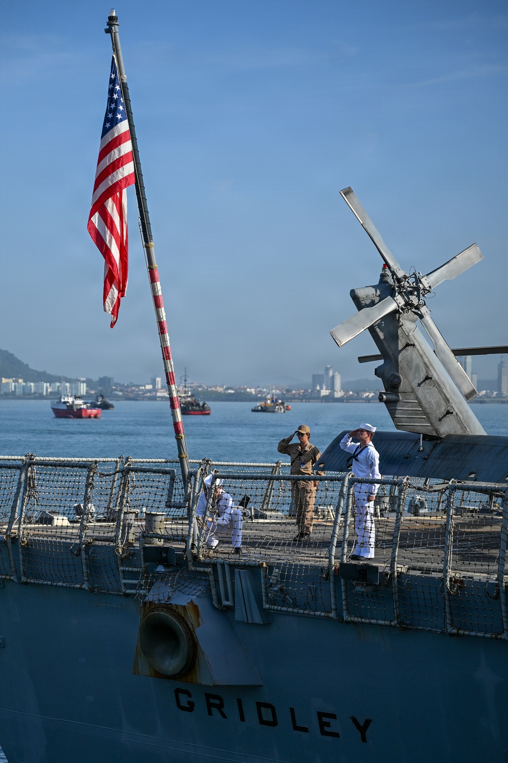 The image shows the USS Gridley, a U.S. Navy vessel, with crew members standing at attention and saluting on the deck. An American flag is prominently displayed on a flagpole at the bow of the ship. The scene appears to take place in a harbor with a coastal cityscape and other vessels visible in the background.