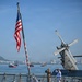 The image shows the USS Gridley, a U.S. Navy vessel, with crew members standing at attention and saluting on the deck. An American flag is prominently displayed on a flagpole at the bow of the ship. The scene appears to take place in a harbor with a coastal cityscape and other vessels visible in the background.