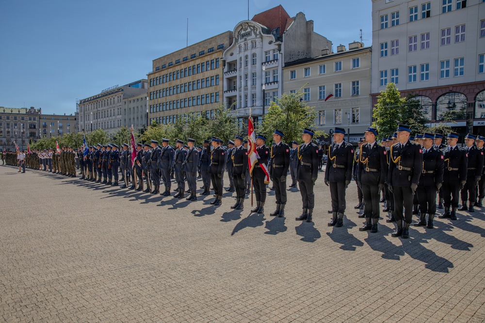 A formal military or police ceremonial parade is taking place in an open public square, likely in Poland, based on the red-and-white flags and architectural style. Personnel in dark navy/blue uniforms with gold belt buckles and peaked caps are standing in formation, with at least one standard-bearer holding a ceremonial flag. The event appears to be an official state or institutional ceremony held on a sunny day.