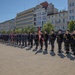A formal military or police ceremonial parade is taking place in an open public square, likely in Poland, based on the red-and-white flags and architectural style. Personnel in dark navy/blue uniforms with gold belt buckles and peaked caps are standing in formation, with at least one standard-bearer holding a ceremonial flag. The event appears to be an official state or institutional ceremony held on a sunny day.