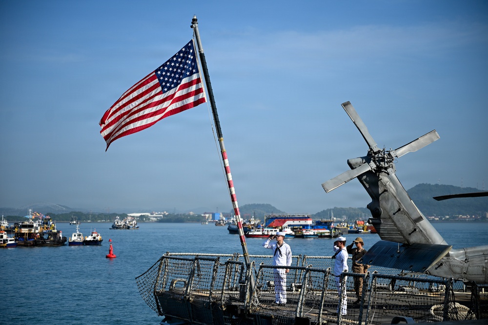 The image shows the stern deck of a U.S. Navy vessel flying a large American flag, with several uniformed personnel visible on deck. A helicopter with folded rotor blades is parked on the right side of the deck. The ship appears to be anchored or moored in a busy harbor with multiple vessels visible in the background.