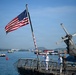 The image shows the stern deck of a U.S. Navy vessel flying a large American flag, with several uniformed personnel visible on deck. A helicopter with folded rotor blades is parked on the right side of the deck. The ship appears to be anchored or moored in a busy harbor with multiple vessels visible in the background.
