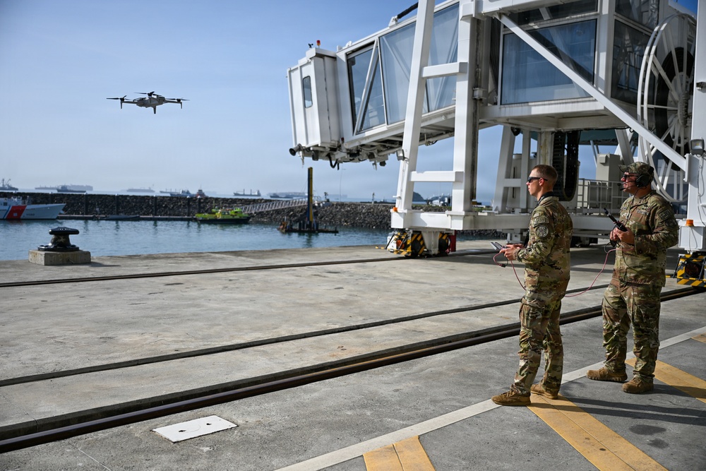 Two military personnel in camouflage uniforms are operating a small quadcopter drone at what appears to be a port or harbor facility. One soldier holds a tablet device while the other holds a remote controller, suggesting a coordinated drone operation. The setting includes a passenger boarding bridge structure, dock infrastructure, and open water with vessels visible in the background.