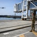 Two military personnel in camouflage uniforms are operating a small quadcopter drone at what appears to be a port or harbor facility. One soldier holds a tablet device while the other holds a remote controller, suggesting a coordinated drone operation. The setting includes a passenger boarding bridge structure, dock infrastructure, and open water with vessels visible in the background.