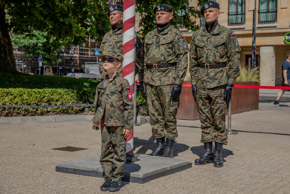 Three adult Polish military soldiers in camouflage uniforms stand at attention near a red-and-white striped flagpole in an outdoor urban setting. A young child dressed in a matching miniature Polish military camouflage uniform and sunglasses stands in front of them, looking upward. The scene appears to be a public military ceremony or event, likely in Poland based on the visible Polish flag patches.