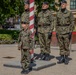 Three adult Polish military soldiers in camouflage uniforms stand at attention near a red-and-white striped flagpole in an outdoor urban setting. A young child dressed in a matching miniature Polish military camouflage uniform and sunglasses stands in front of them, looking upward. The scene appears to be a public military ceremony or event, likely in Poland based on the visible Polish flag patches.
