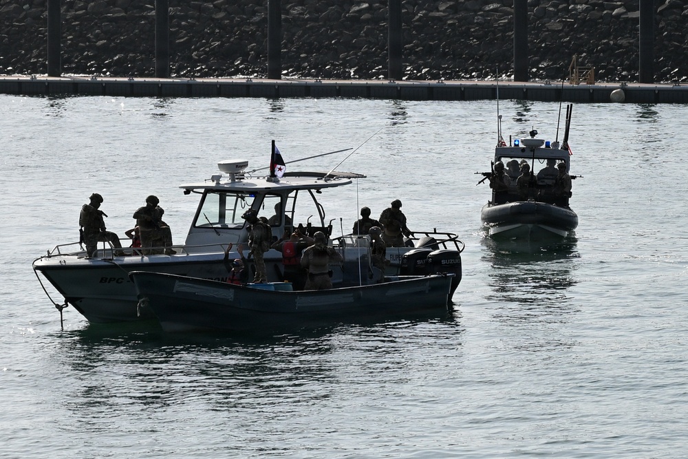 The image shows two military or law enforcement patrol vessels operating in a harbor or coastal waterway. Multiple armed personnel in tactical gear are visible aboard both boats, suggesting a maritime security operation, exercise, or patrol. A flag is visible on the larger vessel, and a rocky breakwater with a dock structure is visible in the background.