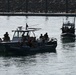 The image shows two military or law enforcement patrol vessels operating in a harbor or coastal waterway. Multiple armed personnel in tactical gear are visible aboard both boats, suggesting a maritime security operation, exercise, or patrol. A flag is visible on the larger vessel, and a rocky breakwater with a dock structure is visible in the background.