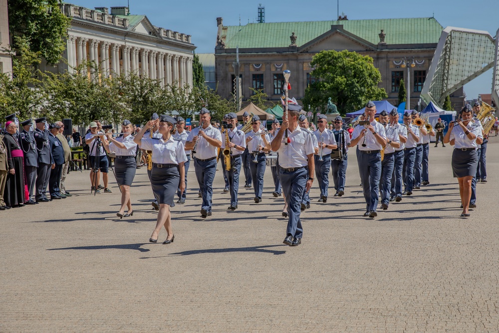 A military marching band performs in formal parade formation on a large public plaza, likely in a Central European city based on the neoclassical architecture visible in the background. The band members wear light blue/grey uniforms with dark trousers or skirts, playing brass and woodwind instruments. Spectators, including what appears to be a Catholic bishop and other dignitaries, observe from the left side.