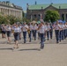 A military marching band performs in formal parade formation on a large public plaza, likely in a Central European city based on the neoclassical architecture visible in the background. The band members wear light blue/grey uniforms with dark trousers or skirts, playing brass and woodwind instruments. Spectators, including what appears to be a Catholic bishop and other dignitaries, observe from the left side.