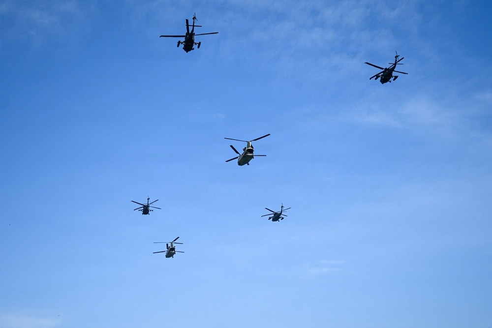 A formation of six military helicopters flying in a loose group against a clear blue sky. The formation includes a mix of what appear to be attack helicopters and tandem-rotor transport helicopters, suggesting a combined arms aerial display or military exercise. The helicopters are captured in silhouette from a ground-level perspective.