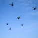A formation of six military helicopters flying in a loose group against a clear blue sky. The formation includes a mix of what appear to be attack helicopters and tandem-rotor transport helicopters, suggesting a combined arms aerial display or military exercise. The helicopters are captured in silhouette from a ground-level perspective.