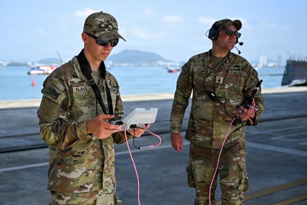 Two U.S. Coast Guard personnel in OCP (Operational Camouflage Pattern) uniforms are operating equipment at what appears to be a port or waterfront facility. The individual in the foreground, identified by name tape as 'KLAUS,' is handling a white device with pink cables, while 'LOCKWOOD' in the background wears a communications headset. Both are wearing sunglasses and their uniforms display U.S. Coast Guard branch tape and American flag patches.